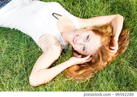 Close up portrait of an attractive young woman laying down on green grass in a park during the summer and listening to music with her earphones, being thoughtful outdoors. 96562280