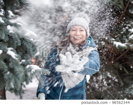 Smiling woman in cable knitted hat is playing with snow. Fun in park between snowy fir trees. Woman laughs as she is throwing snowball. Cold season. Smiling woman in cable knitted hat is playing with snow. Fun in park between snowy fir trees. Woman laughs as she is throwing snowball. Cold season. 96562344