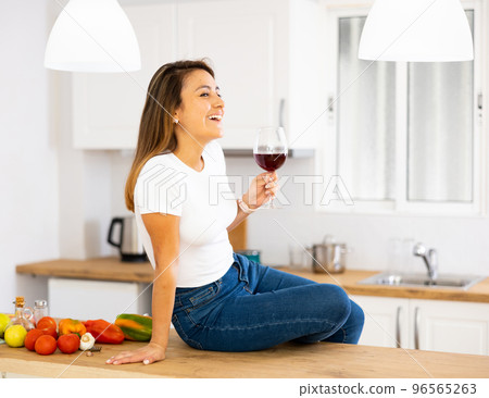 Smiling young Hispanic woman drinking wine sitting on table in kitchen 96565263