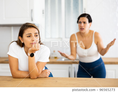 Offended Latina standing in kitchen, listening to disgruntled female friend Offended Latina standing in kitchen, listening to disgruntled female friend 96565273