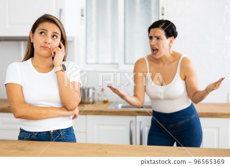 Offended Latina standing in kitchen, listening to disgruntled female friend 96565369