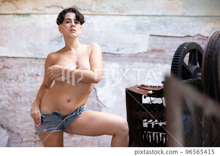Brunette in t-shirt and blue short standing with bare breasts near old rusty machine tools in abandoned plant 96565415