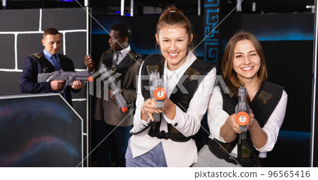 Emotional portrait of two women holding laser pistols and playing laser tag with colleagues Emotional portrait of two women holding laser pistols and playing laser tag with colleagues 96565416