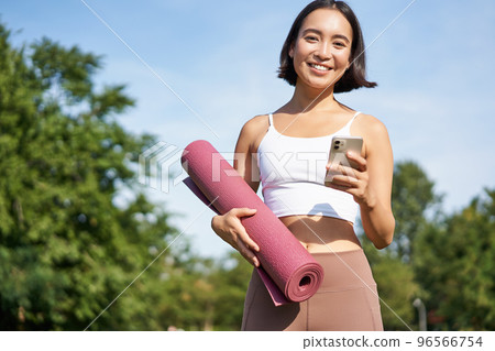 Portrait of young asian woman checking her phone during workout, walking in park with rubber mat, going to the gym, holding smartphone Portrait of young asian woman checking her phone during workout, walking in park with rubber mat, going to the gym, holding smartphone 96566754