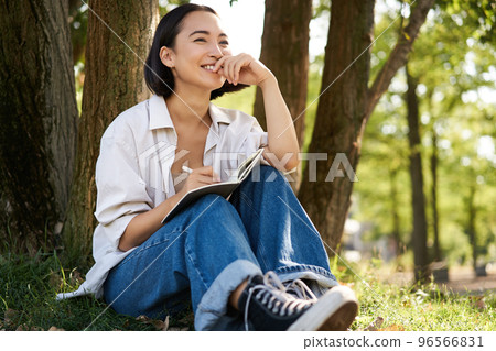 Portrait of young asian woman writing in her notebook, expressing her thoughts on paper in diary, smiling and sitting under tree on summer day in park 96566831