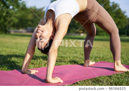Woman smiling while making bridge asana, doing yoga in park on rubber mat 96566835