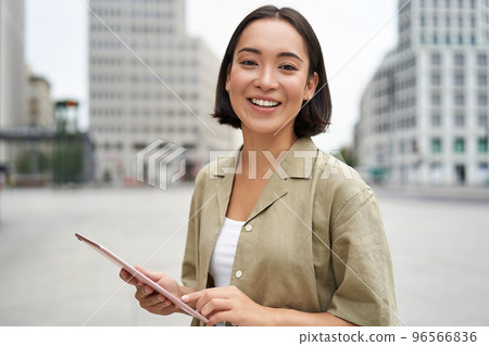 Beautiful girl standing in city square with tablet, reading and drinking takeaway coffee 96566836