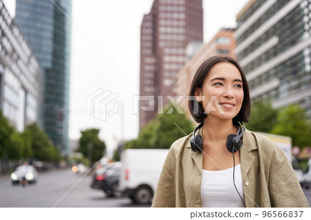 Portrait of young asian woman in headphones, posing in city, smiling and looking away, standing on street of city centre 96566837