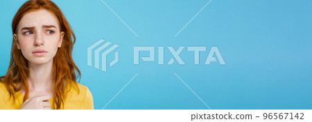 Headshot Portrait of tender redhead teenage girl with serious expression looking at camera. Caucasian woman model with ginger hair posing indoors.Pastel blue background. Copy Space 96567142