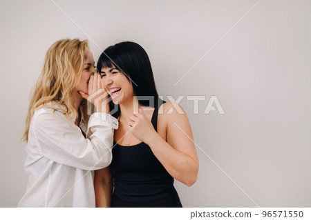 An excited woman in a black swimsuit listens to a friend in a shirt whispering in her ear on a light background An excited woman in a black swimsuit listens to a friend in a shirt whispering in her ear on a light background 96571550