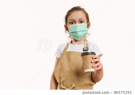 Adorable Caucasian conscious child girl in chef's apron and green protective medical mask, handing out a takeaway coffee tea drink in paper mug to go, looking at camera, isolated over white background Adorable Caucasian conscious child girl in chef's apron and green protective medical mask, handing out a takeaway coffee tea drink in paper mug to go, looking at camera, isolated over white background 96575291
