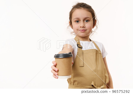 Caucasian lovely little girl wearing a beige chef's apron, handing out on camera a takeaway hot tea cofee drink in cardboard mug, isolated on white, copy space for text. Beverage in paper cup to go Caucasian lovely little girl wearing a beige chef's apron, handing out on camera a takeaway hot tea cofee drink in cardboard mug, isolated on white, copy space for text. Beverage in paper cup to go 96575292