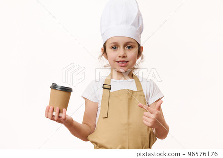 Cute baby girl - barista bartender in white chef hat and beige apron, smiling a beautiful toothy smile looking at camera, pointing at a hot drink in eco paper cup to go, isolated on white background Cute baby girl - barista bartender in white chef hat and beige apron, smiling a beautiful toothy smile looking at camera, pointing at a hot drink in eco paper cup to go, isolated on white background 96576176