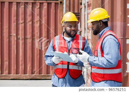 Two short black hair man with moustache and beard dressed in yellow hardhat, red safety vest and white protective glove working during the day under sunlight. There are container in the work area. 96576251