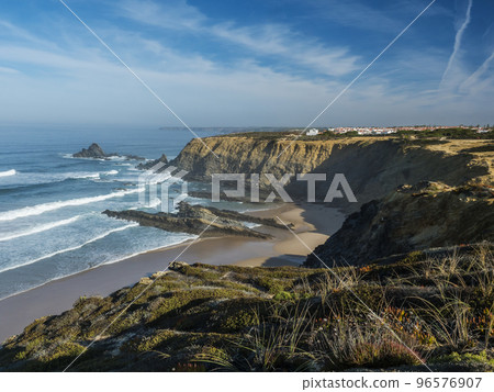 View of golden sand beach and Praia da Zambujeira do Mar village with ocean waves, cliffs and stonesand green vegetation at wild Rota Vicentina coast, Odemira, Portugal. 96576907