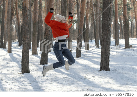 Cheerful young woman jumps high in snowy forest. Walk through the winter wood on sunny day 96577830