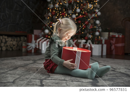 Happy little girl opening xmas gift near Christmas tree. Gift is shine from inside. Cute blonde girl with gift box. 96577831