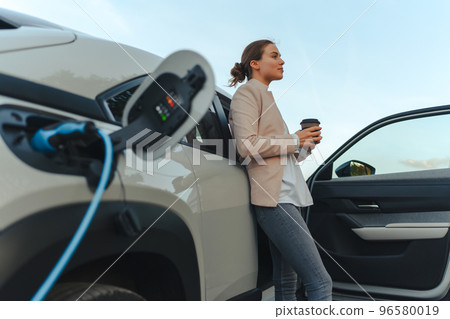 Young woman with cup of coffee waiting while her electric car charging, sustainable and economic transportation concept. 96580019