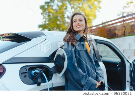 Young woman waiting while her electric car charging in home charging station, sustainable and economic transportation concept. 96580051