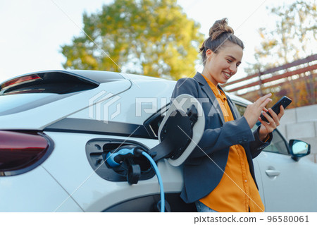 Young woman with smartphone waiting while her electric car charging in home charging station, sustainable and economic transportation concept. 96580061