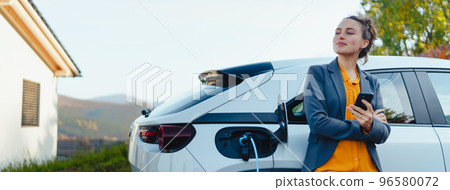 Young woman with smartphone waiting while her electric car charging in home charging station, sustainable and economic transportation concept. Young woman with smartphone waiting while her electric car charging in home charging station, sustainable and economic transportation concept. 96580072