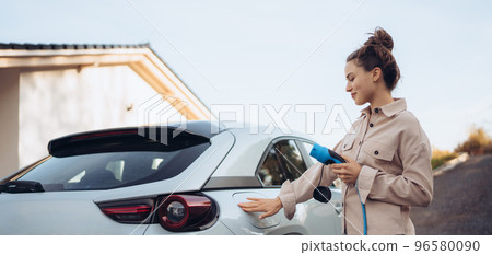 Young woman holding power supply cable from her electric car, prepared for charging it in home, sustainable and economic transportation concept. Young woman holding power supply cable from her electric car, prepared for charging it in home, sustainable and economic transportation concept. 96580090