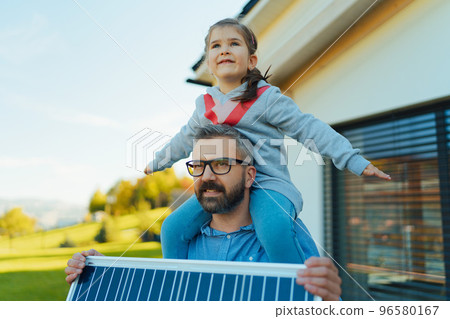 Father with his little daughter on piggyback, catching sun at solar panel,charging it at their backyard. Alternative energy, saving resources and sustainable lifestyle concept. 96580167