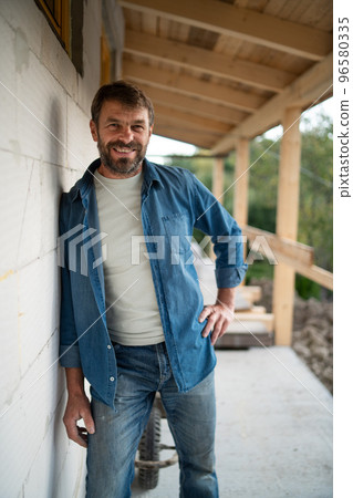 Happy mature man standing in front of his new house under construction, looking at camera. 96580335