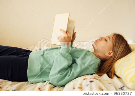 Caucasian blond teenage girl is lying on the bed and reading book. 96580727