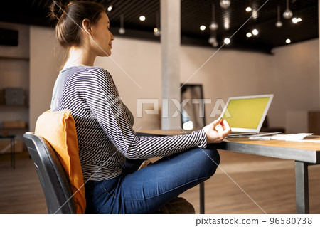 Brunette woman freelancer sitting in lotus pose and meditating with hands in mudra gesture on chair 96580738