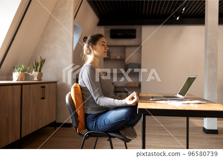 Brunette woman freelancer sitting in lotus pose and meditating with hands in mudra gesture on chair 96580739
