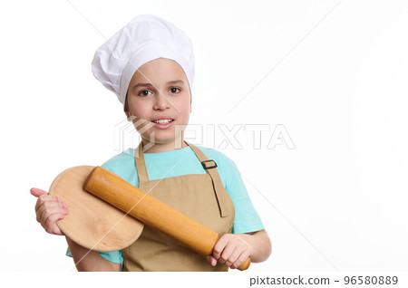 Multi-ethnic teenage boy - baker wearing chef uniform, smiles a cheerful toothy smile, posing to camera with a wooden cutting board and rolling pin in his hands, isolated over white background Multi-ethnic teenage boy - baker wearing chef uniform, smiles a cheerful toothy smile, posing to camera with a wooden cutting board and rolling pin in his hands, isolated over white background 96580889