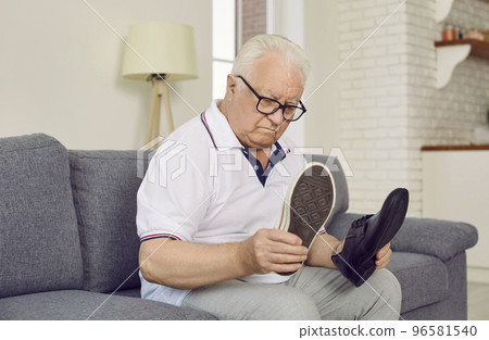 Demented retired senior man sitting on couch and choosing which type of shoes to wear 96581540