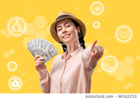 A happy smiling young woman holds a fan of dollars and points her finger forward. Yellow background with digital infographics. The concept of cryptocurrency and apps for trading on the stock exchange A happy smiling young woman holds a fan of dollars and points her finger forward. Yellow background with digital infographics. The concept of cryptocurrency and apps for trading on the stock exchange 96582250