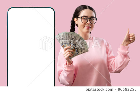 Investment App. Big smartphone with white screen and smiling caucasian woman holds a fan of dollar bills and thumb up at pink background. Mock up. Concept of cash back and template of finance Investment App. Big smartphone with white screen and smiling caucasian woman holds a fan of dollar bills and thumb up at pink background. Mock up. Concept of cash back and template of finance 96582253