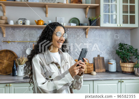 A young beautiful Latin American woman is standing in the kitchen at home, using a telephone. Smiling, she types messages, reads the news, makes phone calls. A young beautiful Latin American woman is standing in the kitchen at home, using a telephone. Smiling, she types messages, reads the news, makes phone calls. 96582355