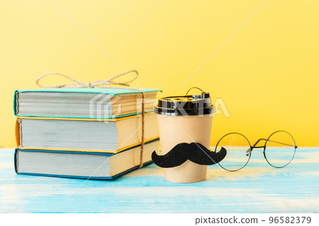Cup of coffee with a felt moustache on the background of a stack of books and glasses. Copy space. Concept of Father's day 96582379