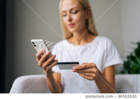 Low-angle view of beautiful blonde housewife typing card number on mobile phone at home. Close-up of pretty blonde female looking smartphone screen indoors. Focused lady making payment by cellphone. Low-angle view of beautiful blonde housewife typing card number on mobile phone at home. Close-up of pretty blonde female looking smartphone screen indoors. Focused lady making payment by cellphone. 96582630