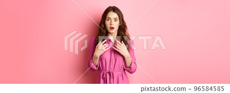 Shocked young feminine woman gasping, holding hands on chest and staring speechless at camera, look with disbelief, standing against pink background 96584585