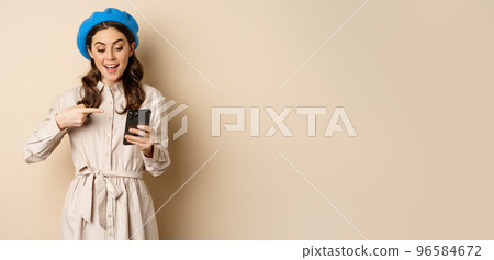 Portrait of stylish modern woman in outerwear, pointing at mobile phone screen and looking happy, smiling pleased, standing over beige background 96584672