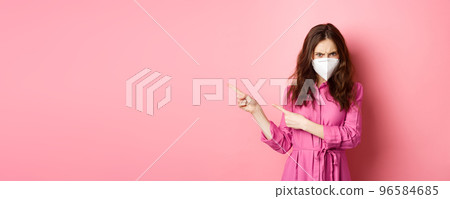 Covid-19, quarantine and social distancing concept. Angry woman frowning and looking judgemental, wearing face mask respirator, pointing fingers left, pink background 96584685