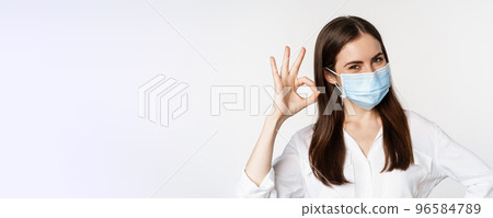 Close up portrait of smiling female office worker in medical mask, woman showing okay sign, excellent sign, using personal protection from covid pandemic, white background 96584789