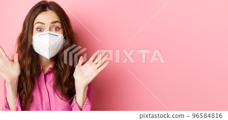 Coronavirus, preventive measures and health concept. Close up portrait of surprised and excited woman in respirator, raising hands up and gasping amazed, pink background 96584816