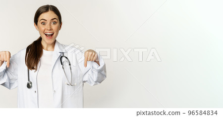Portrait of happy female doctor, pointing fingers down and smiling, demonstrating promo offer, discount, standing over white background Portrait of happy female doctor, pointing fingers down and smiling, demonstrating promo offer, discount, standing over white background 96584834