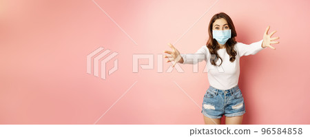 Portrait of smiling friendly girl in medical face mask, reaching hands, stretching arms forward for hug, receiving smth, standing over pink background Portrait of smiling friendly girl in medical face mask, reaching hands, stretching arms forward for hug, receiving smth, standing over pink background 96584858