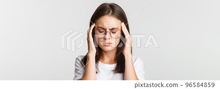 Close-up of young woman in glasses touching head and grimacing from pain, having headache or migraine, feeling sick Close-up of young woman in glasses touching head and grimacing from pain, having headache or migraine, feeling sick 96584859