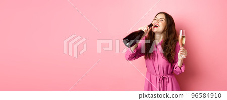 Celebration and holidays concept. Happy carefree woman drinking champagne from bottle with pleased smile, relaxing on party, holding glass, standing over pink background 96584910