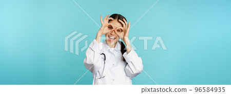 Funny therapist, girl doctor showing okay, binoculars zero gesture on eyes and smiling, fooling around and having fun, standing over blue background Funny therapist, girl doctor showing okay, binoculars zero gesture on eyes and smiling, fooling around and having fun, standing over blue background 96584935