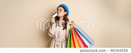 Portrait of cute happy girl kissing her credit card after buying gifts, holding shopping bags, standing over beige background Portrait of cute happy girl kissing her credit card after buying gifts, holding shopping bags, standing over beige background 96584936