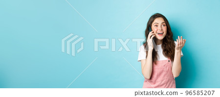 Portrait of cheerful modern girl talking on phone, gesturing and looking happy, smiling at camera, chat with friend on smartphone, standing over blue background Portrait of cheerful modern girl talking on phone, gesturing and looking happy, smiling at camera, chat with friend on smartphone, standing over blue background 96585207